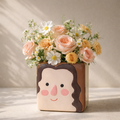 Floral arrangement in a vase shaped like a face with a smiley face design on a neutral background