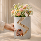 Cylindrical concrete vase with decorative elements and a hand holding it, against a neutral background.